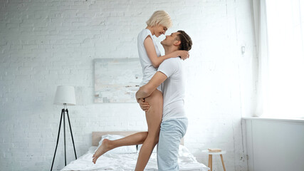Fototapeta na wymiar side view of cheerful man holding in arms blonde woman in bedroom
