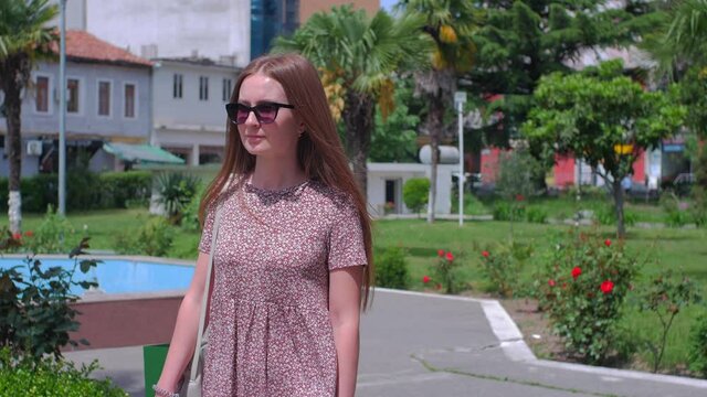 Young redhead caucasian woman in sunglasses walks in the green garden near palms