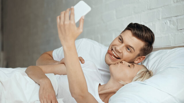 young woman kissing man sticking out tongue while taking selfie on smartphone