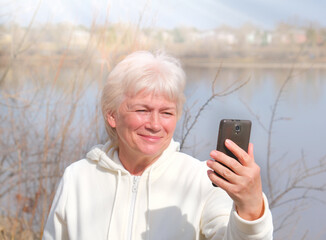 Portrait of happy blonde senior woman holding camera and taking selfie. Selective focus, Focus on phone