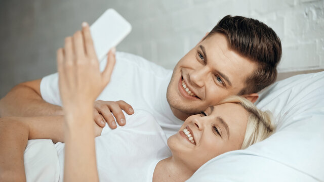 happy young man and woman taking selfie on smartphone in bedroom