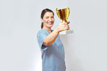 Middle aged nurse in a light blue scrub smiling proudly and holding a golden trophy up, white...