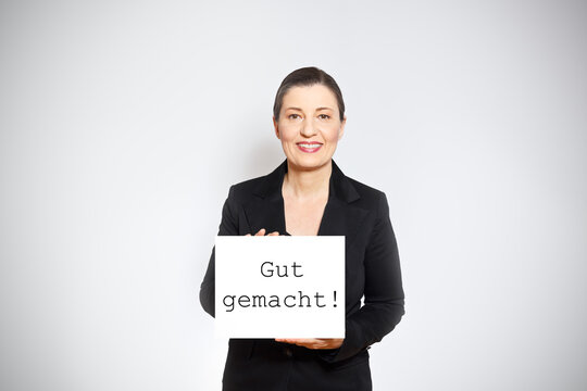 Smiling Middle Aged Woman In Black Blazer Holding Up A Sign With The Text Well Done In German, White Background.