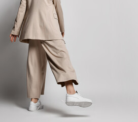 Legs of woman in beige business smart casual pantsuit, sneakers walking over light background with copy space. Back view. Stylish business female wear and fashion concept on gray background