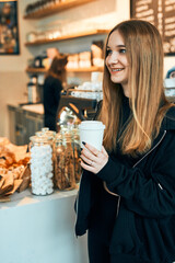 Woman holding coffee cup, standing in a coffee shop and waiting for the order. Smiling young woman having a break drinking coffee in cafe