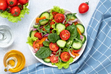 Summer vegetable salad with cherry tomatoes, cucumbers, arugula, basil and seasoned with olive oil. Blue checkered napkin on a gray table. View from above. Healthy diet, vegetarian food close-up.