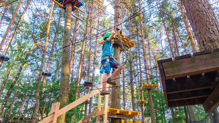 A boy in a helmet climbs a rope ladder stretched between trees in a high-rope adventure park. Family sports leisure with children. Types of extreme sports activities for children during the holidays.