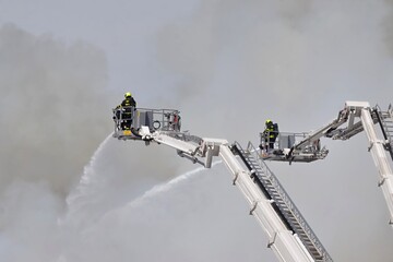 Firefighters on a high-altitude aerial platform battling a huge scrap yard fire with massive smoke...