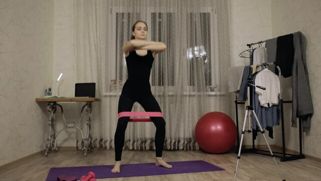 Athletic woman in sportswear doing squats with resistance band while training at home 
