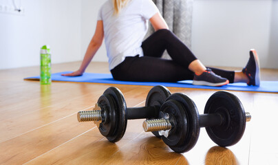 Fototapeta na wymiar Home workout. Unrecognizable blonde woman in black leggings and sneakers doing stretching after strength training with dumbbells Woman on a pink mat at home. Focus on the dumbbells in the foreground.