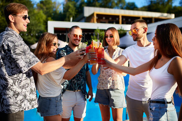 Group of friends having fun at poolside summer party, clinking glasses with colorful summer cocktails outdoors near hotel swimming pool. People toast drinking fresh juice at luxury villa on vacation.