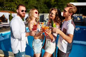 Group of friends having fun at poolside summer party, clinking glasses with colorful summer cocktails outdoors near hotel swimming pool. People toast drinking fresh juice at luxury villa on vacation.