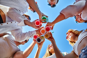 Low angle view of friends having fun at poolside summer party, clinking glasses with colorful...