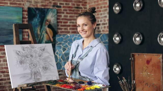 Portrait of female artist mixing color paints with art brush on the palette. Young beautiful painter at the studio preparing for painting a still life picture with sunflowers.