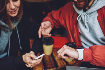 Cropped male and female hipsters checking time on synchronized smartphone and cellular technology during meeting in sidewalk cafe, millennial bloggers reading notification on digital devices