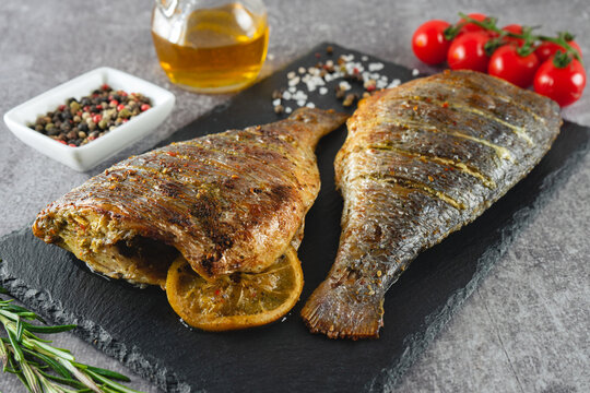Grilled fish with roasted with lemon, rosemary, tomatoes, olive oil and spices on black slate dish, on grey background.