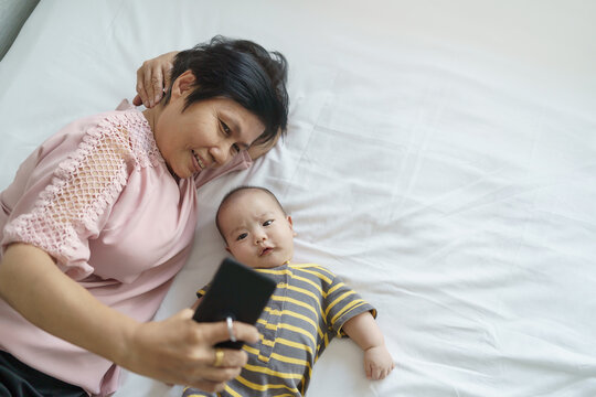 Happy Asian Grandmother and little baby boy or Grandson lying on bed taking selfie photos and doing video call with mobile phone or smartphone
