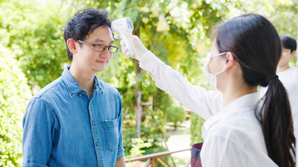 Young Asia female restaurant staff wearing protective face mask using infrared thermometer checker or temperature gun on customer's forehead before enter. Lifestyle new normal after corona virus.