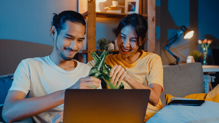 Happy young asian couple enjoy night party event online sit couch use laptop computer video call with friends toast drink beer via video call online in living room at home, Social distancing concept.