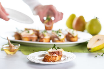 Close up of a pear and prosciutto crostini with hands taking one from a platter of them in behind, in soft focus.