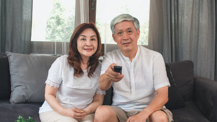 Obraz premium Asian elderly couple watching television in living room at home, sweet couple enjoy love moment while lying on the sofa when relaxed at home. Enjoying time lifestyle senior family at home concept.