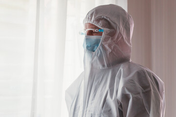Fototapeta premium Young Caucasian men in hazmat suit wearing surgical face mask and face shield during Coronavirus pandemic prevention. Young physician doctor in protective ppe suit standing by the window
