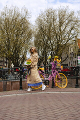 Fototapeta premium A beautiful young blonde woman walking along the canal and a red bike with a bouquet of yellow tulip flowers in a long blue dress and brown coat. Street Style in Amsterdam. Travel concept.