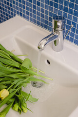 A bouquet of fresh yellow tulips with green stems in a sink with water running from the faucet in a modern bathroom decorated with blue tile.