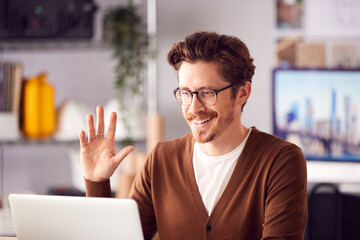 Male Architect In Office Making Video Call At Desk On Laptop Waving At Screen