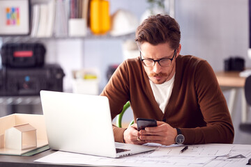 Male Architect In Office With Laptop Working On Plan At Desk Text Messaging On Mobile Phone