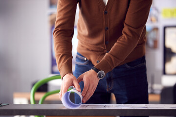 Close Up Of Male Architect In Office Unrolling Building Plan Or Blueprint On Desk