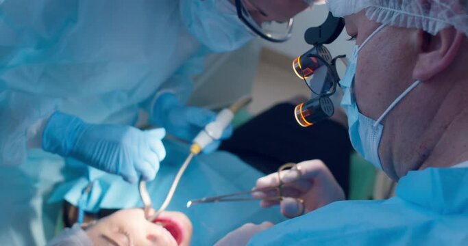 A male dentist is wearing a shadowless headlamp on the head is doing female patient teeth treatment by using forceps with the surgical thread from a mouth and a nurse is holding a saliva ejector.
