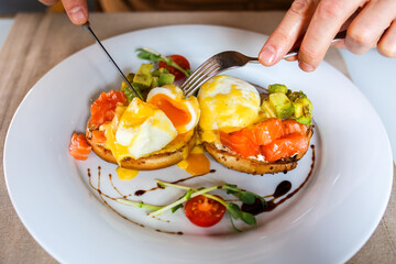 Eggs Benedict. The poached egg is cut with a knife. Breakfast plate. Eggs benedicts on toast, close-up.
Delicious breakfast with egg Benedict. The egg is spreading. Man eating breakfast with knife and