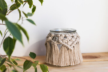 Beautiful jar candle holder with macrame decor on wood shelf. Boho home decor. Houseplant in front. Rustic decoration. - obrazy, fototapety, plakaty