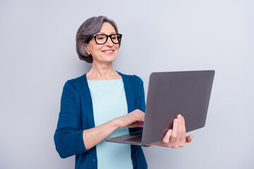 Portrait of attractive cheerful gray haired woman holding in hands using laptop typing isolated over grey pastel color background