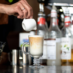 Making latte coffee. Barista pours freshly brewed coffee into whipped milk froth in front of bottles on blurred background. Glass of coffee drink with froth. Close up shot.