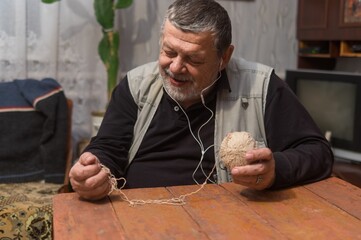 Indoor portrait of positive senior Ukrainian peasant taking clew of thread while sitting at old...