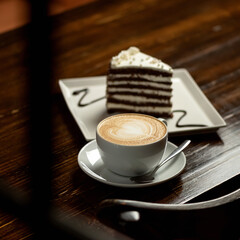 Cup of creamy cappuccino coffee. Portion of hot aromatic drink with slice of cake on dark wooden table. Square format, soft focus. Shot through the window. Copy space.