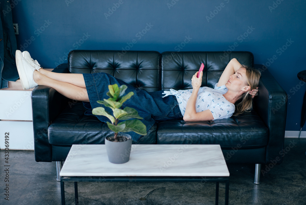 Wall mural Young woman lying on the couch is resting and looking at the phone social networks.