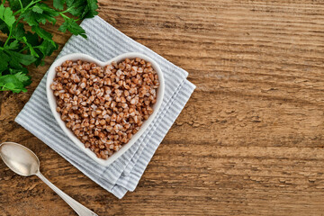 Heart-shaped bowl of boiled buckwheat porridge on old wooden background. Gluten free ancient grain for healthy diet. Top view. Copy space.