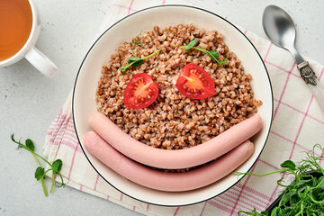 Boiled buckwheat porridge, meat sausages, tomato and microgreens of peas in a white plate in stone grey background. Fun food art idea healthy lunch for kids. Top view.
