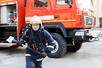 Portrait of serious and confident caucasian fireman standing and holding hammer, wearing special protective uniform in the truck background