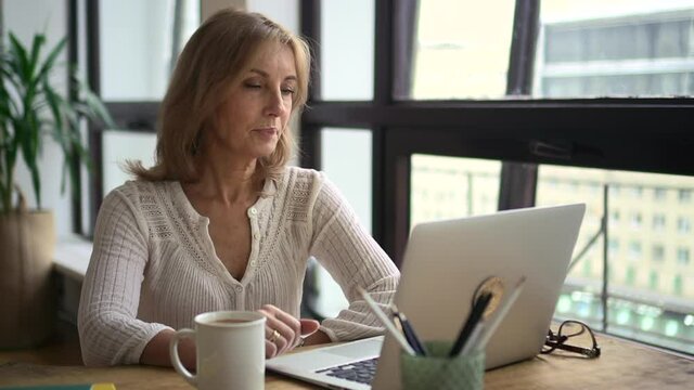 Woman has online talk in front of laptop screen and sits at table in home office.