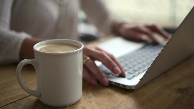 Closeup view of woman working with laptop and sitting at table with coffee in home office.