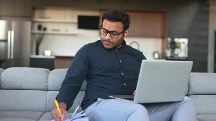 Busy multi-ethnic hindu freelancer guy using laptop computer for work or studying on the distance from home, concentrated indian man watching online webinar and taking notes in notebook, e-learning - Powered by Adobe