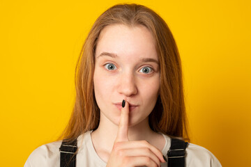 Fototapeta premium Serious young girl dressed casually holding finger at her lips asking not to make noise or hold tongue and conceal some private confidential information. Top secret.