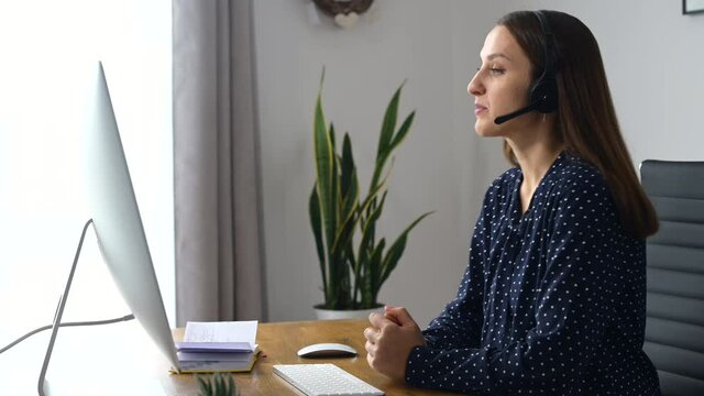 Friendly and cheerful woman wearing headset takes a call, using trendy PC for video connection with customer or colleagues on the distance, a female with hands free device at the workplace