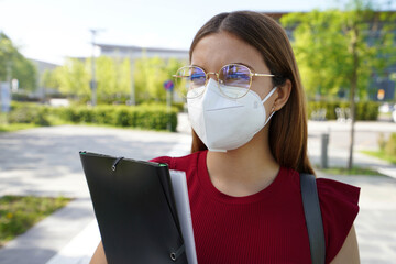 Female university student with FFP2 KN95 face mask walking in city street. College girl back to school holding bag and folders.