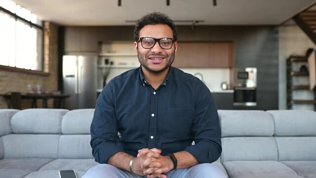 Video chat with indian male employee, mixed race colleague. Screen with a mixed-race guy on it, a man in eyeglasses and shirt sits on the couch looks at camera and waving, video meeting, video call