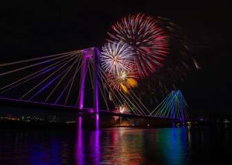 Festive fireworks in honor of Victory Day on Great Patriotic War in Krasnoyarsk, Russia. Bright flashes of fireworks in the dark sky above the colorful suspension bridge with water reflection